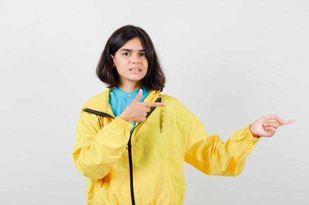 Teen girl in shirt, yellow jacket pointing right and looking cheerless , front view.