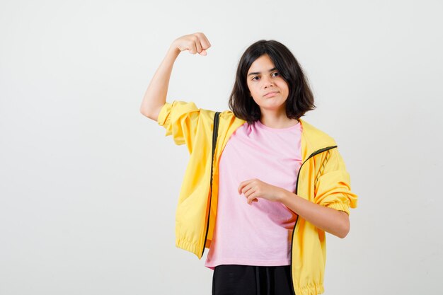 Девушка-подросток показывает мышцы рук в футболке, куртке и смотрит сосредоточенно, вид спереди.