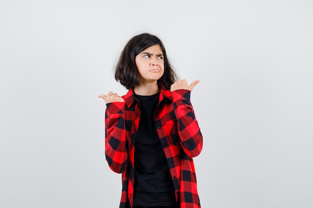 Teen girl showing both sides with thumbs in t-shirt, checkered shirt and looking indecisive. front view