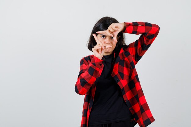 Teen girl showing frame gesture in t-shirt, checkered shirt and looking confident , front view.