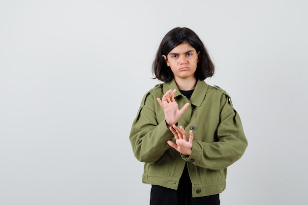 Teen girl showing stop gesture in army green jacket and looking gloomy. front view.