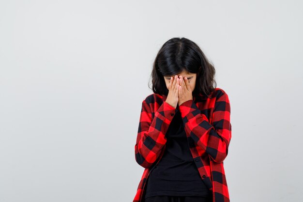 Teen girl in t-shirt, checkered shirt holding hands on face and looking sorrowful , front view.