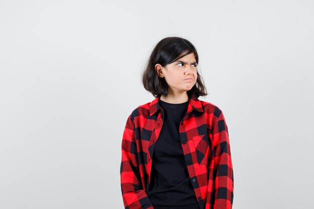 Teen girl in t-shirt, checkered shirt looking aside and looking spiteful , front view.