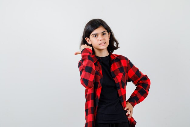 Teen girl in t-shirt, checkered shirt pointing to the left side with thumb and looking cheery , front view.