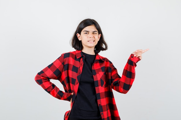 Teen girl in t-shirt, checkered shirt pointing to the right side and looking pleased , front view.