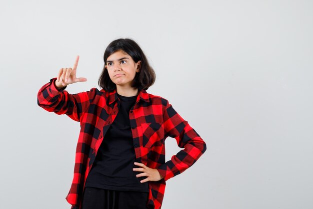 Teen girl in t-shirt, checkered shirt pointing up and looking careful , front view.