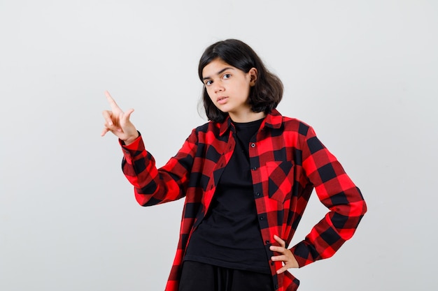 Teen girl in t-shirt, checkered shirt pointing at upper left corner and looking careful , front view.