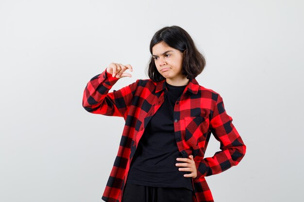 Teen girl in t-shirt, checkered shirt showing small size sign and looking dissatisfied , front view.