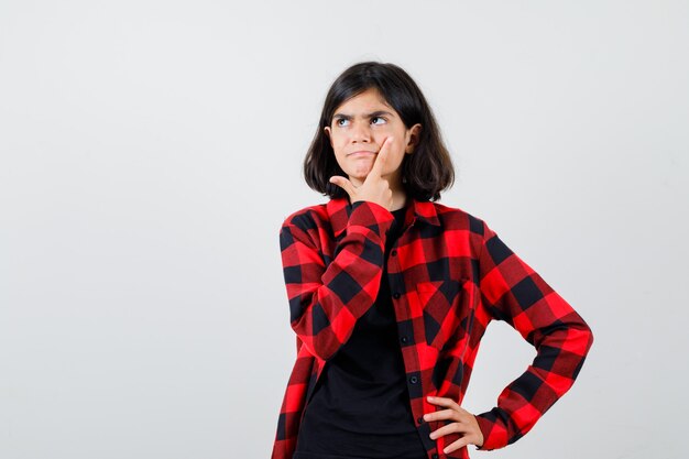 Teen girl in t-shirt, checkered shirt standing in thinking pose and looking pensive , front view.