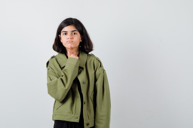 Teen girl in t-shirt, green jacket holding finger on chin and looking careful , front view.