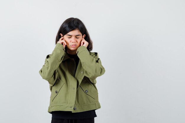 Teen girl in t-shirt, green jacket plugging ears with fingers and looking stressed , front view.