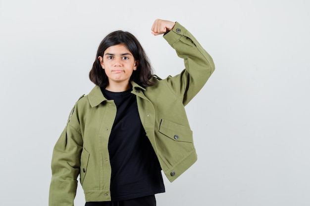 Teen girl in t-shirt, green jacket showing muscles of arm and looking confident , front view.
