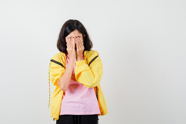 Teen girl in yellow tracksuit, t-shirt holding hands on face and looking upset , front view.