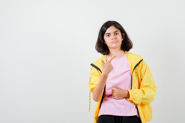 Teen girl in yellow tracksuit, t-shirt pointing up and looking dissatisfied , front view.