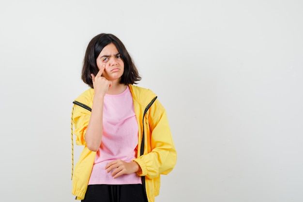 Teen girl in yellow tracksuit, t-shirt pulling eye down with finger and looking sad , front view.