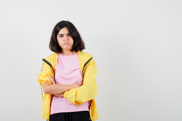 Девушка-подросток в желтом спортивном костюме, футболка стоит со скрещенными руками, надувает щеки и выглядит недовольной, вид спереди.