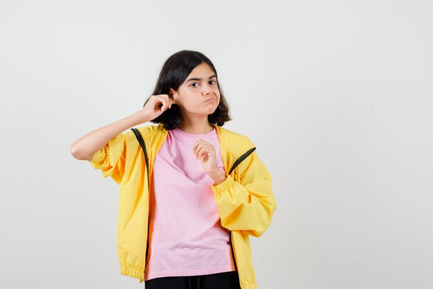 Teen girl in yellow tracksuit, t-shirt standing with pulling ear down and looking displeased , front view.