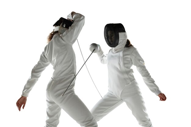 Teen girls in fencing costumes with swords in hands isolated on white studio background. Young female models practicing and training in motion, action. Copyspace. Sport, youth, healthy lifestyle.