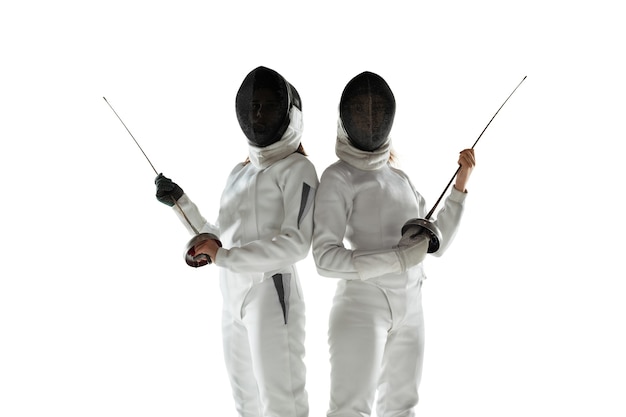 Free photo teen girls in fencing costumes with swords in hands isolated on white studio background. young female models practicing and training in motion, action. copyspace. sport, youth, healthy lifestyle.