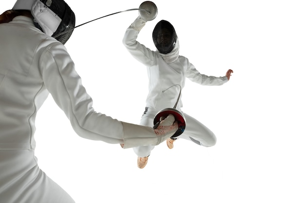 Teen girls in fencing costumes with swords in hands isolated on white studio background. Young female models practicing and training in motion, action. Copyspace. Sport, youth, healthy lifestyle.