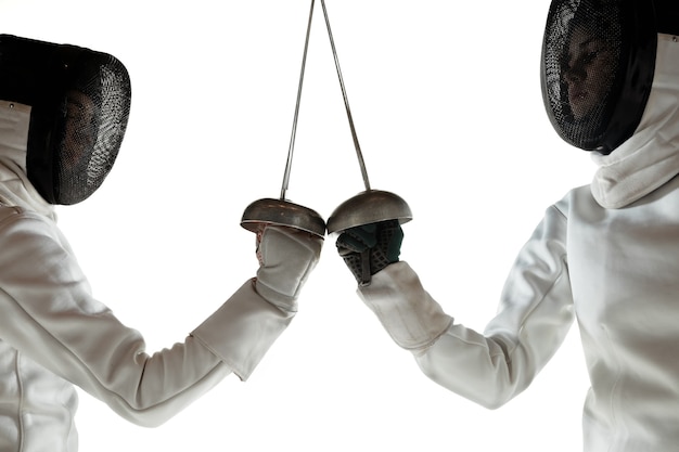 Teen girls in fencing costumes with swords in hands isolated on white studio background. Young female models practicing and training in motion, action. Copyspace. Sport, youth, healthy lifestyle.