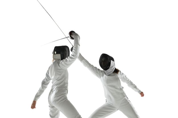 Teen girls in fencing costumes with swords in hands isolated on white studio background. Young female models practicing and training in motion, action. Copyspace.