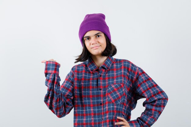 Teen woman in checked shirt and beanie spreading palm aside looking cheery