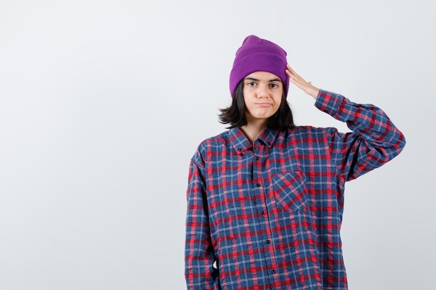 Девушка-подросток в клетчатой рубашке и фиолетовой шапочке показывает радостный жест приветствия