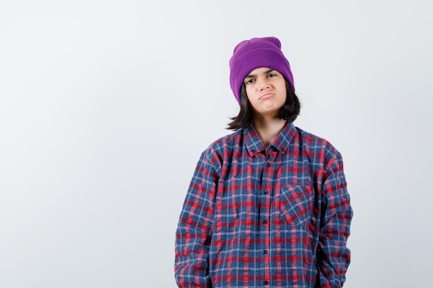 Free photo teen woman in checkered shirt and beanie curving lips looking dissatisfied