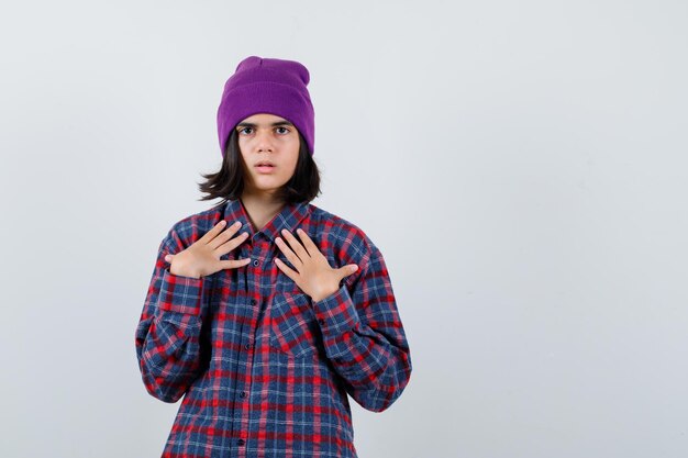 Teen woman in checkered shirt and beanie holding hands on chest looking focused