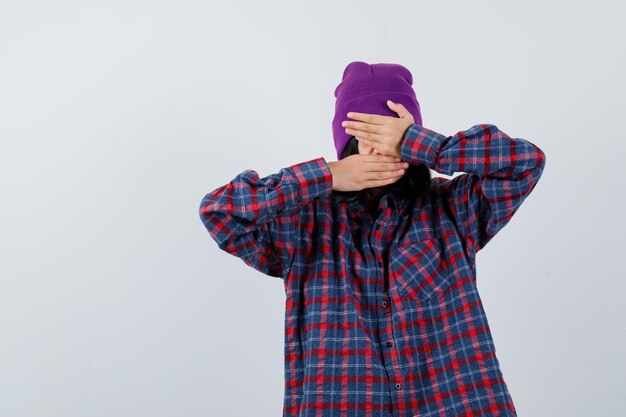 Teen woman in checkered shirt and beanie holding hands on face looking unwell