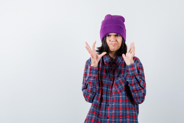 Teen woman in checkered shirt and beanie keeping hands in puzzled manner looking annoyed