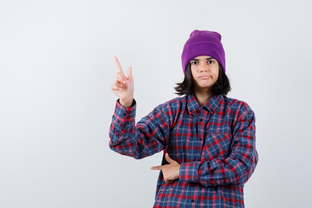 Teen woman in checkered shirt and beanie pointing up and looking careful