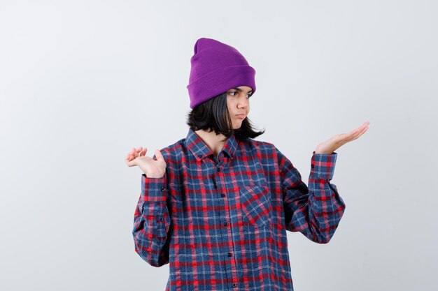 Teen woman in checkered shirt and beanie pretending to show something looking indecisive