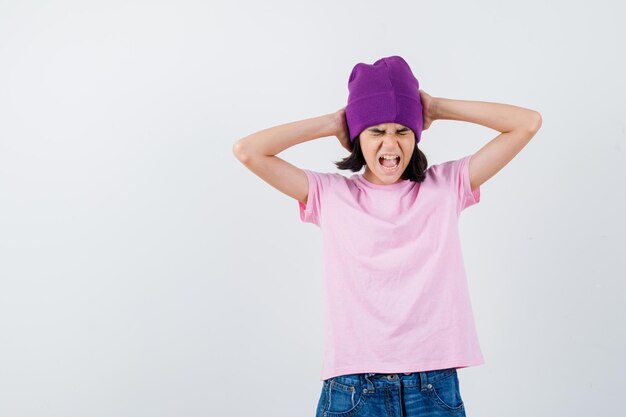 Teen woman clasping head with hands while screaming in t-shirt and beanie looking anxious