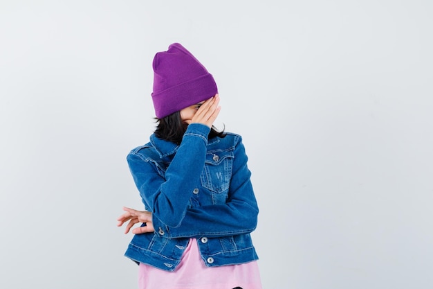 Молодая женщина в джинсовой куртке-бини, пряча лицо за рукой, смущенно смотрит