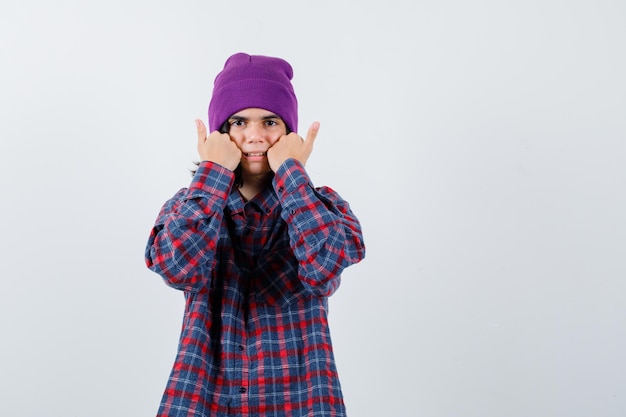 Foto gratuita teen donna che tiene i pugni sulle guance in camicia a scacchi e berretto guardando attento