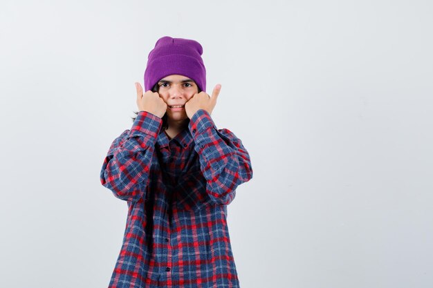 Teen woman holding fists on cheeks in checkered shirt and beanie looking careful