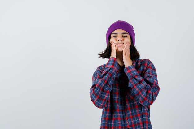 Teen woman holding hands on cheeks in checked shirt purple beanie looking cute 