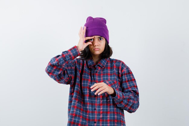 Teen woman opening eye with fingers in checkered shirt and beanie