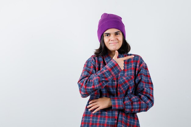 Teen woman pointing right with index finger in checked shirt and beanie looking cheery