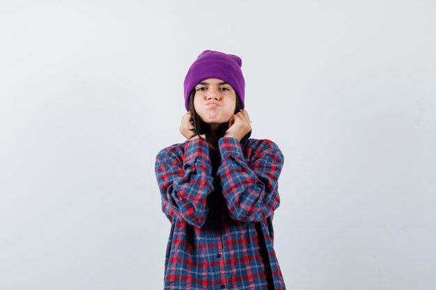 Teen woman pulling down her earlobes while blowing cheeks in checkered shirt