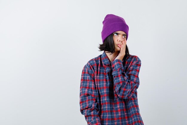 Teen woman pulling down her eyelid in checkered shirt and beanie and looking bored