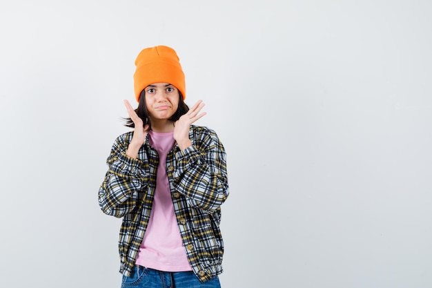 Teen woman raising hands near face in t-shirt jacket beanie looking excited