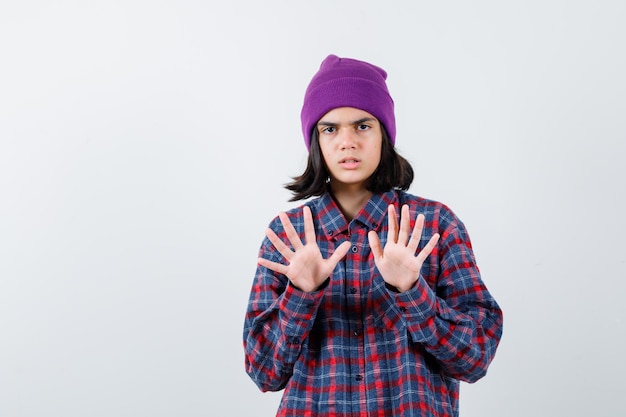 Teen woman raising palms to stop in checked shirt purple beanie looking scared