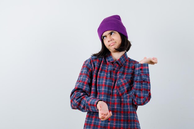 Teen woman spreading palms aside in checkered shirt and beanie looking dissatisfied