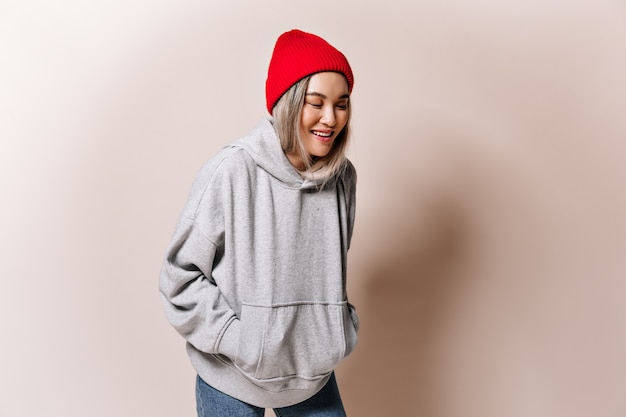 Teen woman in sweatshirt and hat laughs on beige wall