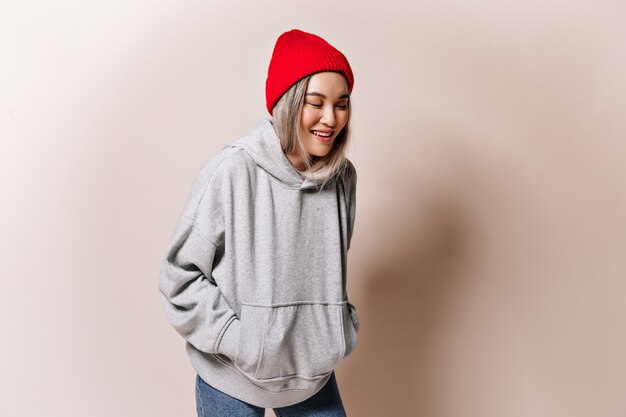 Teen woman in sweatshirt and hat laughs on beige wall