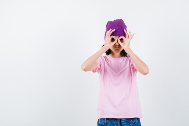 Teen woman in t-shirt and beanie showing glasses gesture looking curious