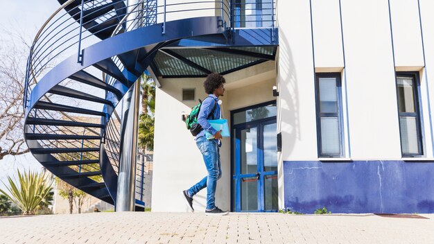 Teenage boy holding book in hand walking in front of university building